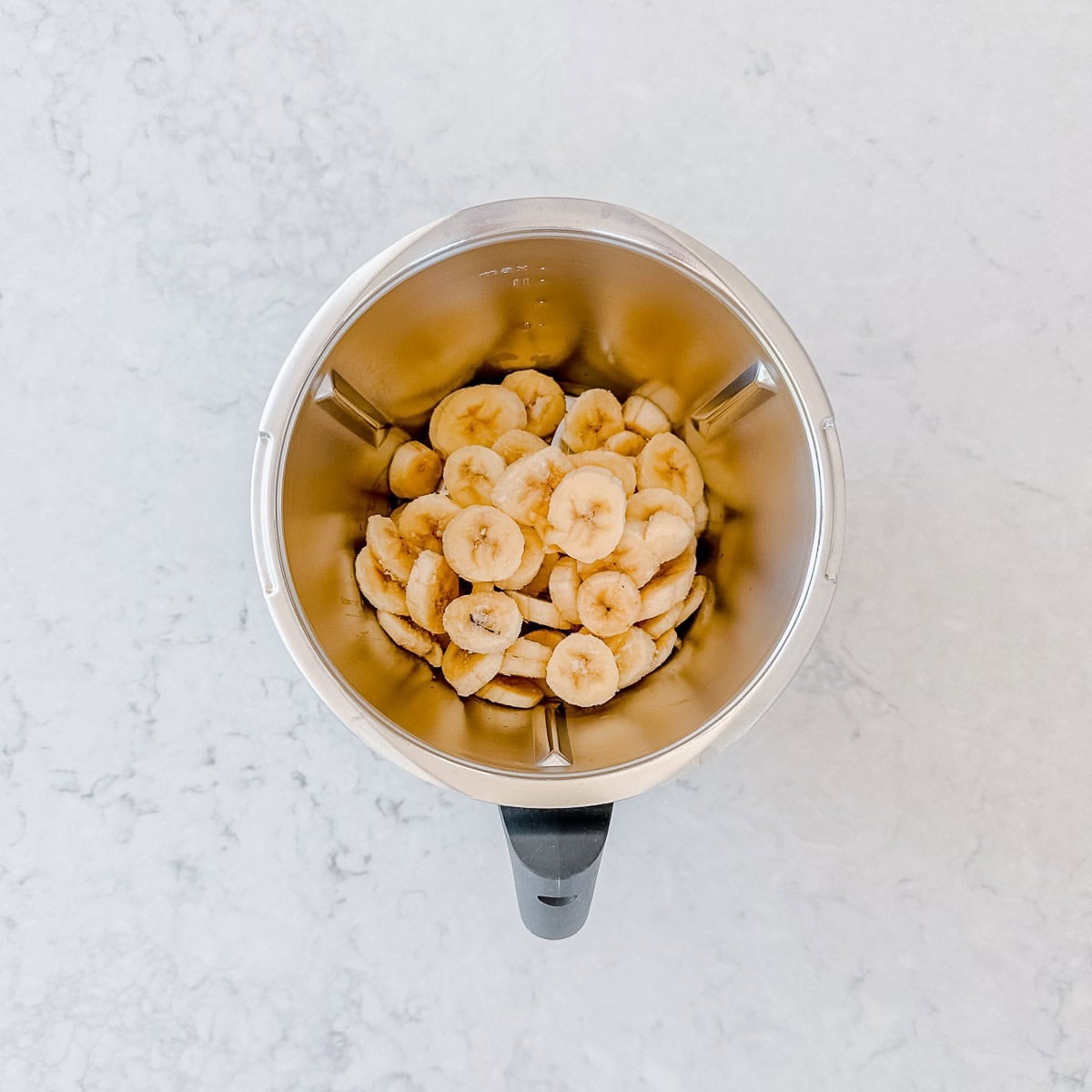 A top-down view of a Thermomix jug filled with sliced bananas, placed on a light-coloured, marble-like surface.