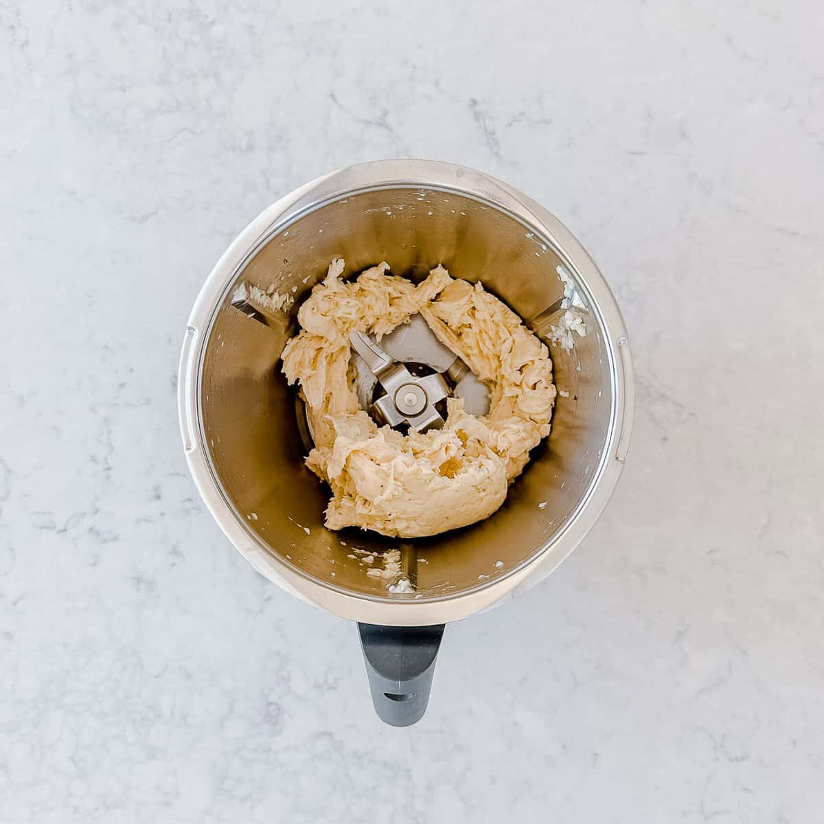 A top-down view of a Thermomix bowl containing blended frozen bananas, sitting on a light-coloured, marbled worktop.