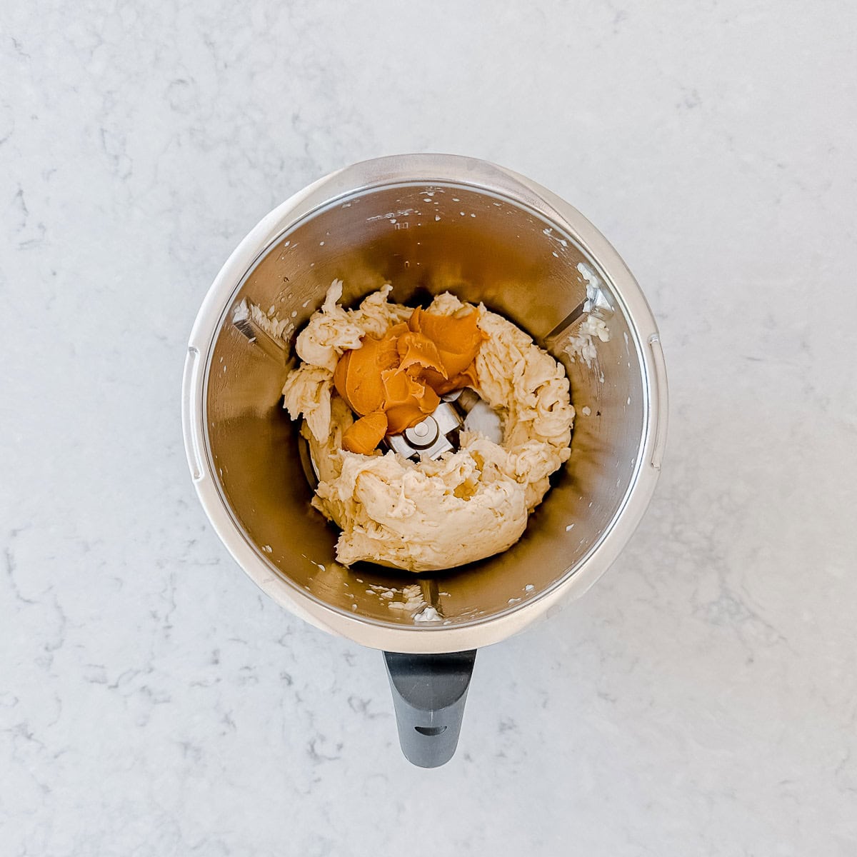 A top-down view of a Thermomix bowl containing blended frozen bananas and peanut butter, sitting on a light-coloured, marbled worktop.