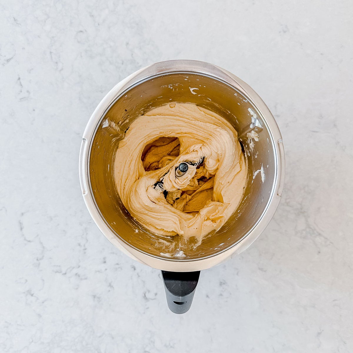Overhead view of creamy banana ice cream being mixed in a metal bowl with a black handle, set on a light, marbled worktop.