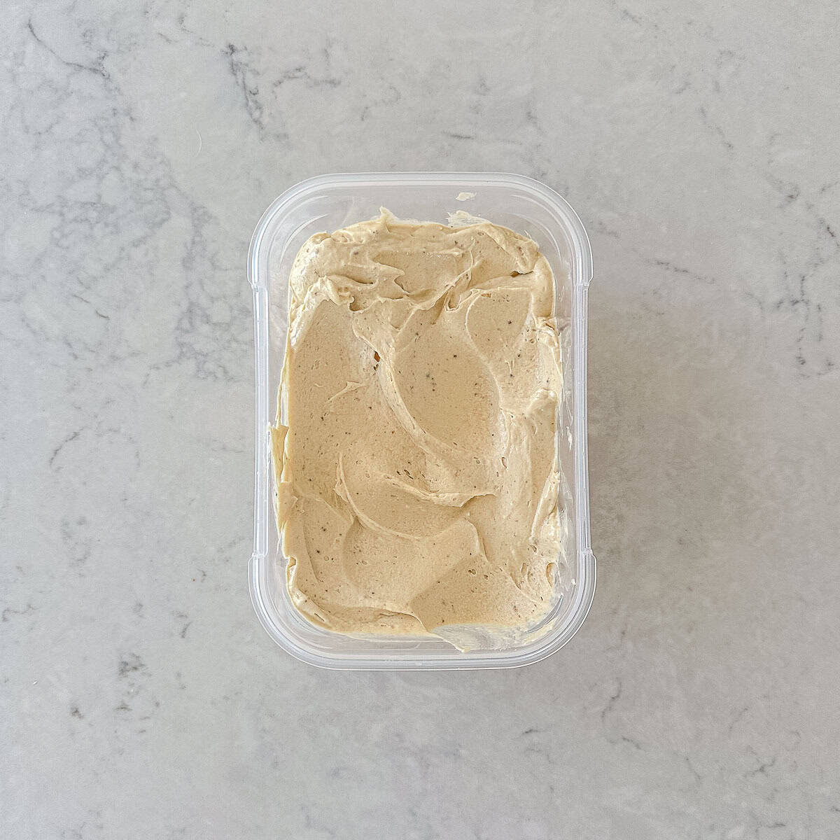 A clear plastic container filled with smooth, light banana ice cream sits on a light grey marbled surface. The ice cream has a swirled, whipped texture on top.