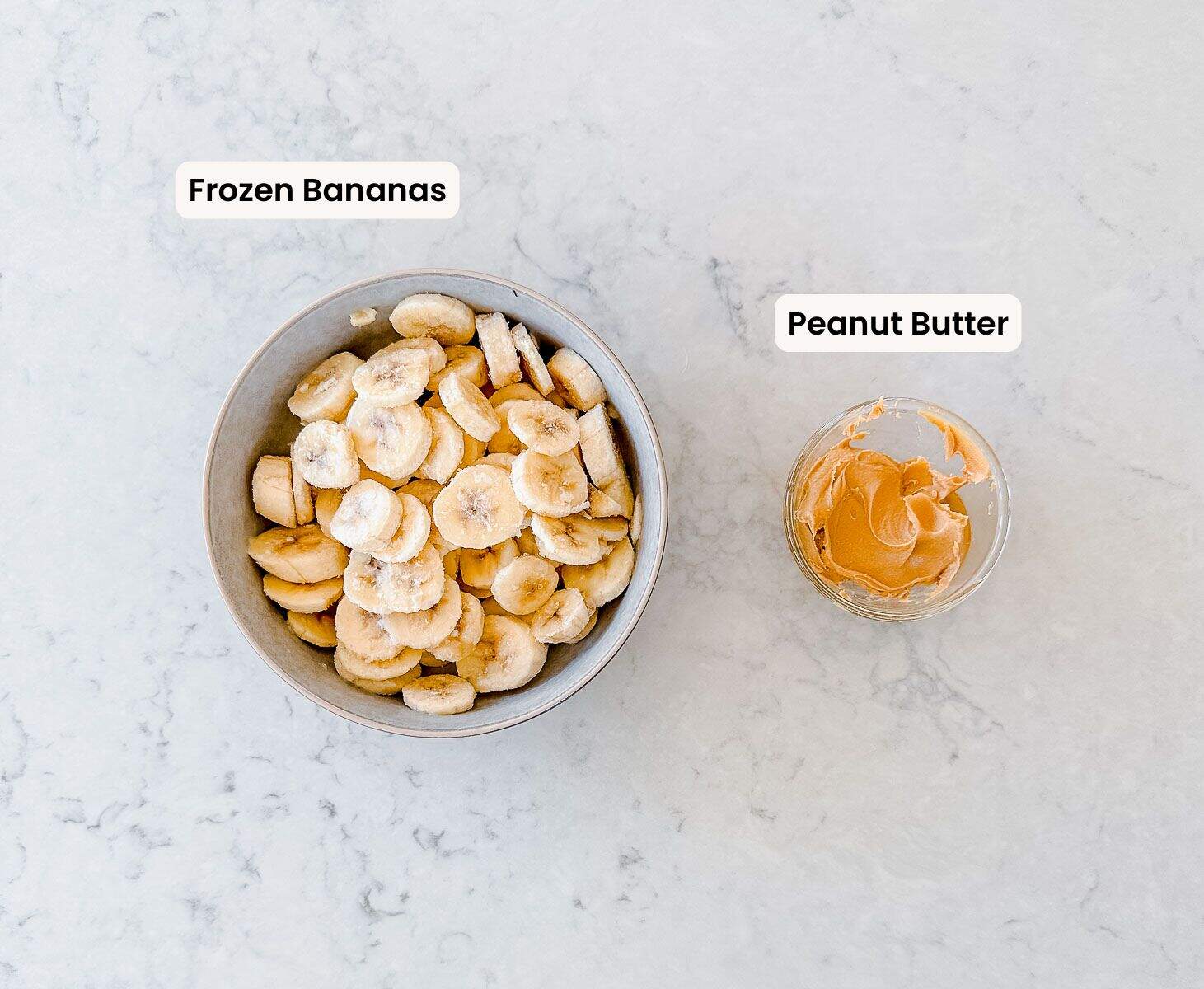 A bowl of sliced frozen bananas is on the left, labelled Frozen Bananas. To the right is a small dish of peanut butter, labelled Peanut Butter, both on a light marble surface.