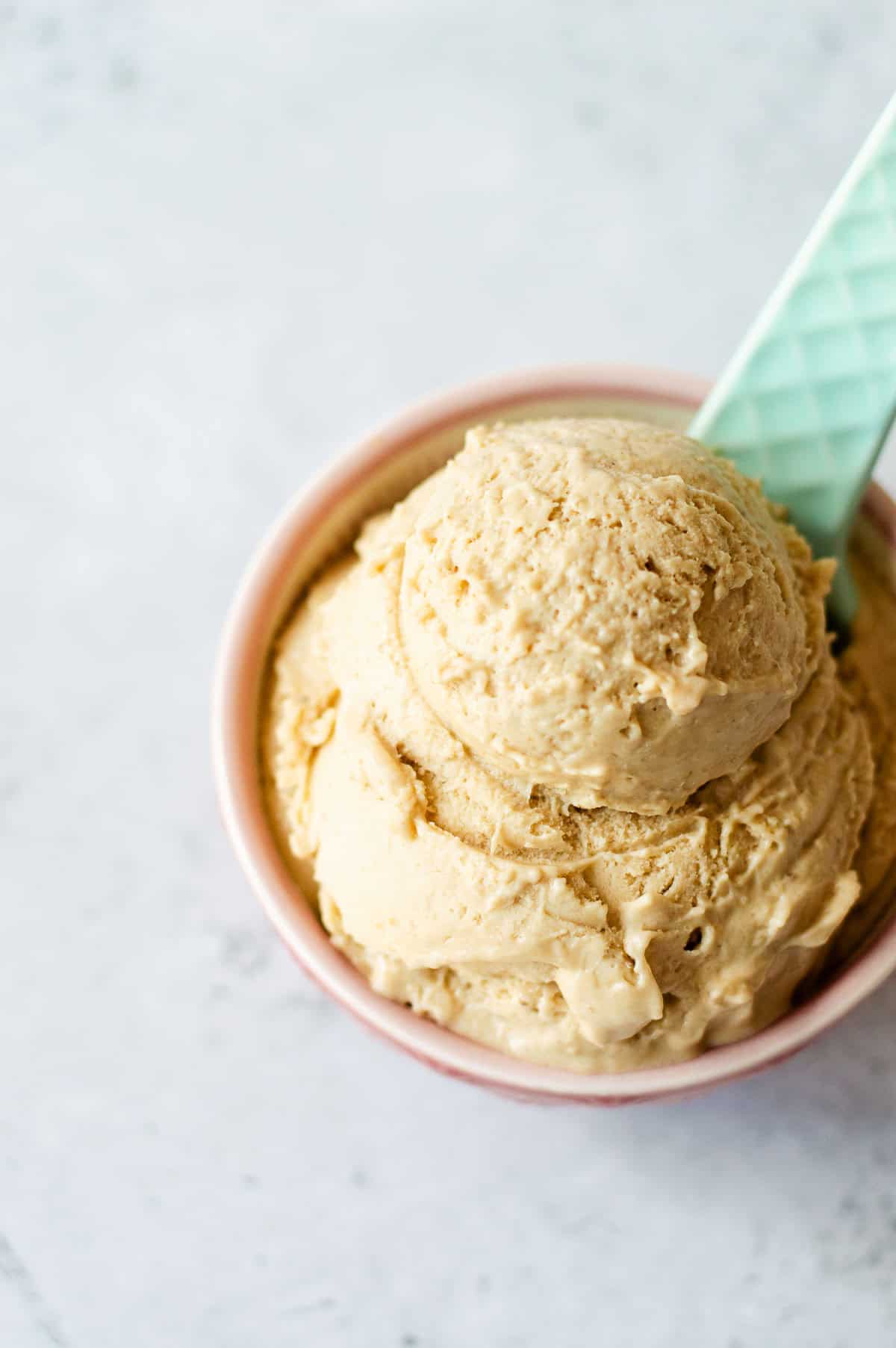 A bowl of creamy brown ice cream with two scoops, served in a pink bowl with a light blue textured spoon on a light marble surface.