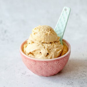 A pink bowl with white star patterns holds two scoops of light brown ice cream, with a pale blue spoon resting in the bowl. The background is a light, textured surface.