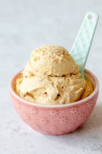 A pink bowl with white star patterns holds two scoops of light brown ice cream, with a pale blue spoon resting in the bowl. The background is a light, textured surface.