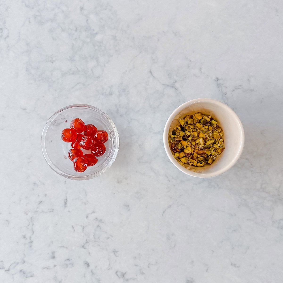 Two small bowls on a white marble surface: one contains bright red cherries, and the other holds chopped green pistachios.