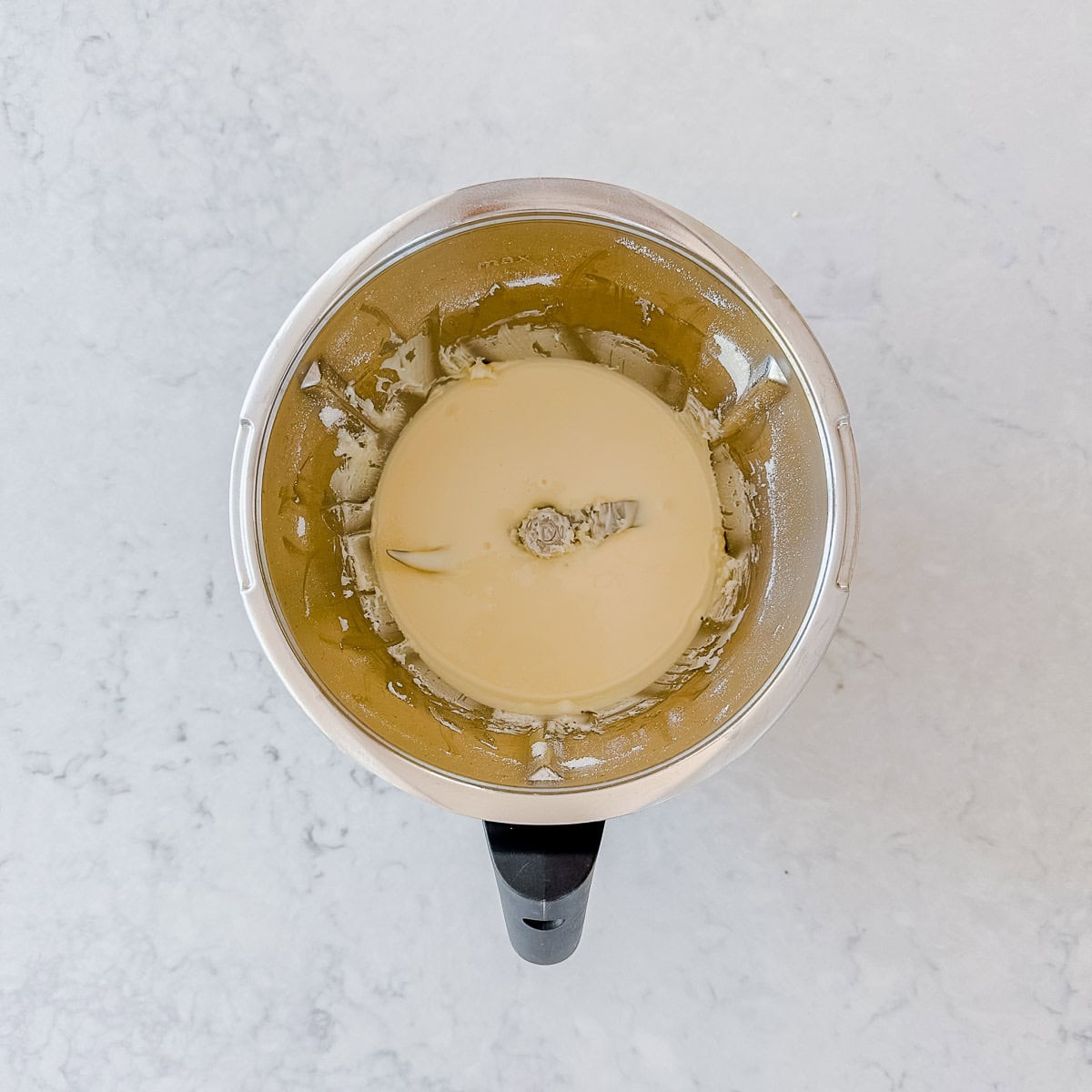 A top-down view of a metal mixing bowl with a creamy, light-colored mixture and a central mixing blade, placed on a white marble surface.