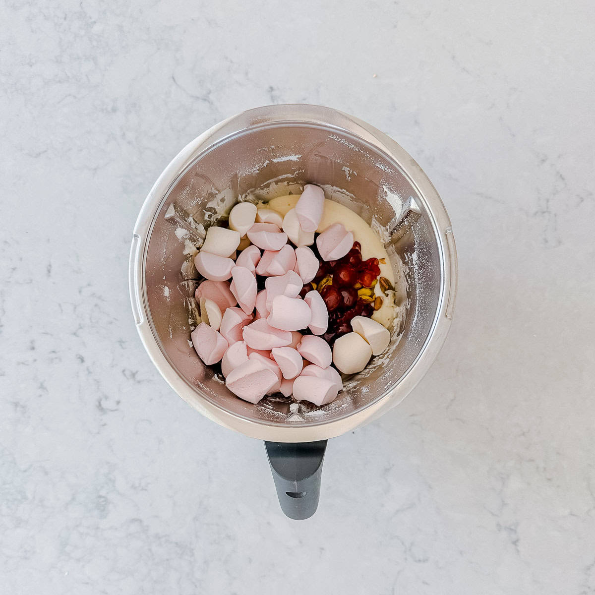 A mixing bowl with pink marshmallows, red and brown ingredients, and a creamy mixture inside, placed on a light marble worktop.