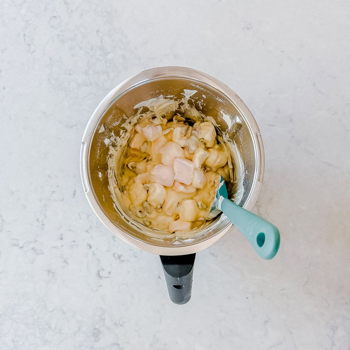 A mixing bowl filled with chopped ingredients and a creamy mixture, being stirred with a blue spatula, sits on a white marble surface.