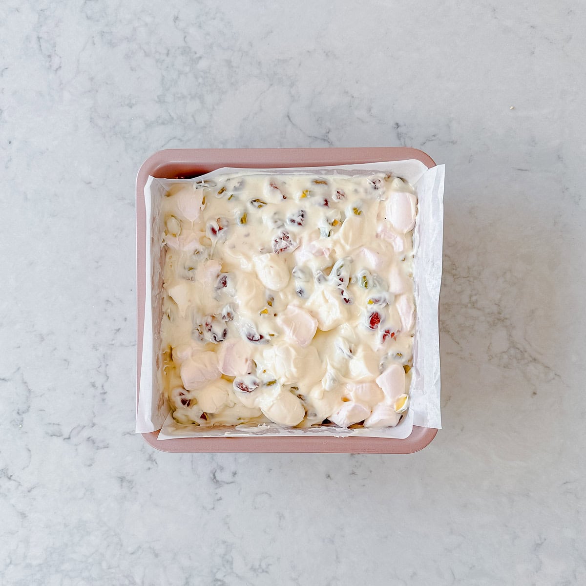 A square baking pan lined with parchment paper, filled with a mixture of white chocolate, marshmallows, nuts, and dried fruit, sits on a light marble surface.