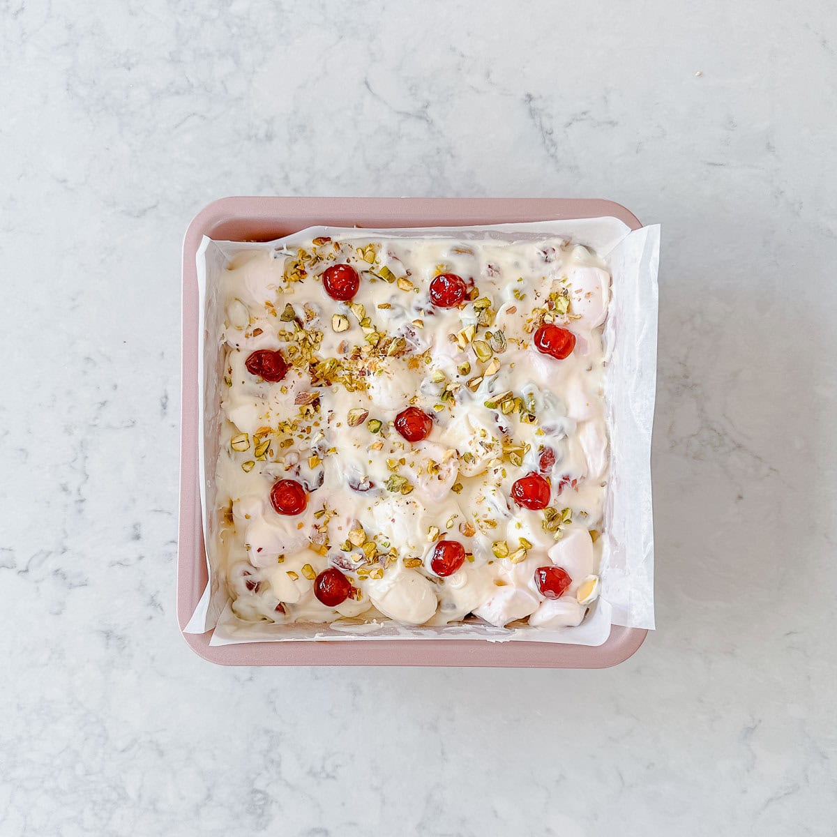 A square baking pan lined with parchment paper, filled with a creamy white dessert topped with red cherries and chopped pistachios, on a light marble surface.