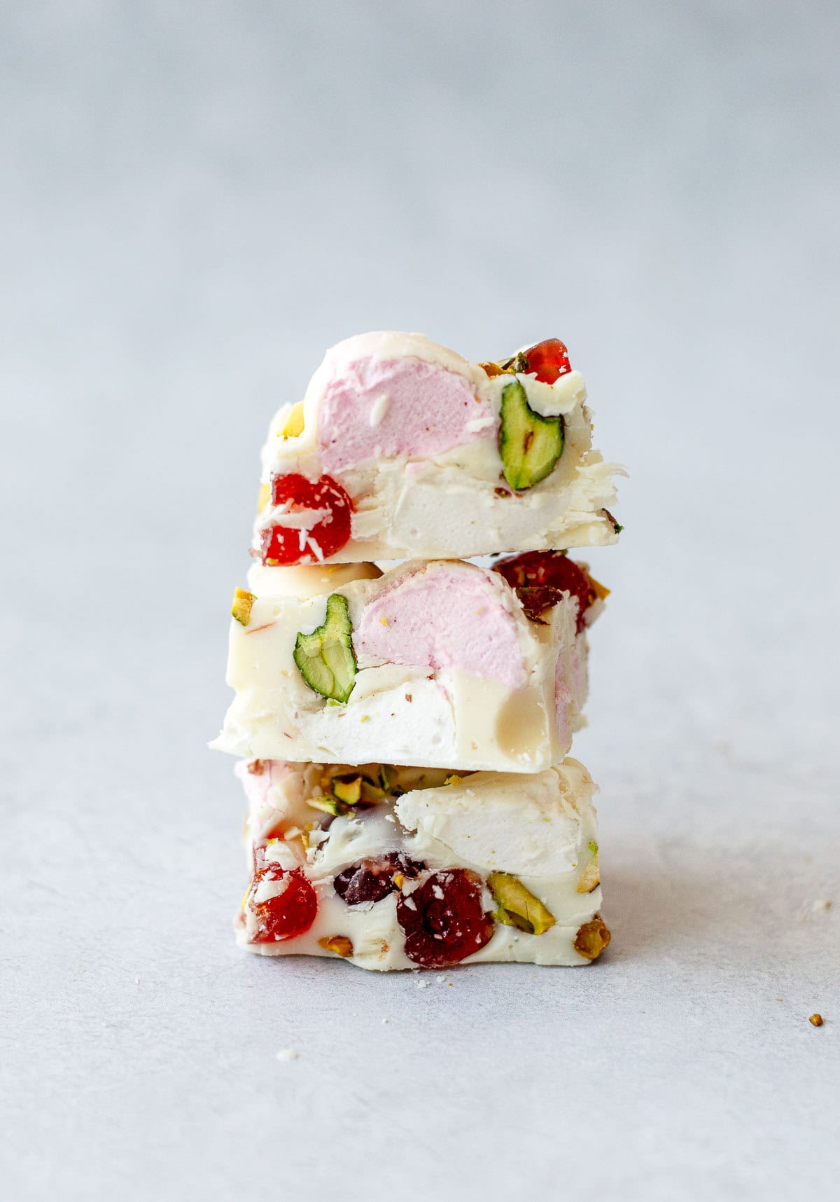 Three pieces of white chocolate rocky road stacked on a light surface, filled with green pistachios, red dried fruit, and pink marshmallows.