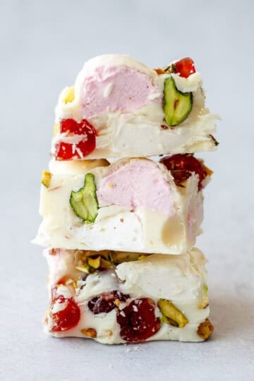 Three pieces of white rocky road with visible pistachios, red glacé cherries, and pink marshmallow are stacked on a light grey surface.