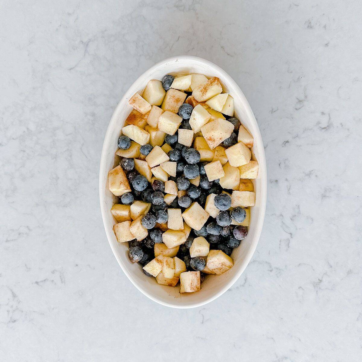 Oval white dish filled with chopped apples and blueberries, sprinkled with cinnamon, on a light marble surface.
