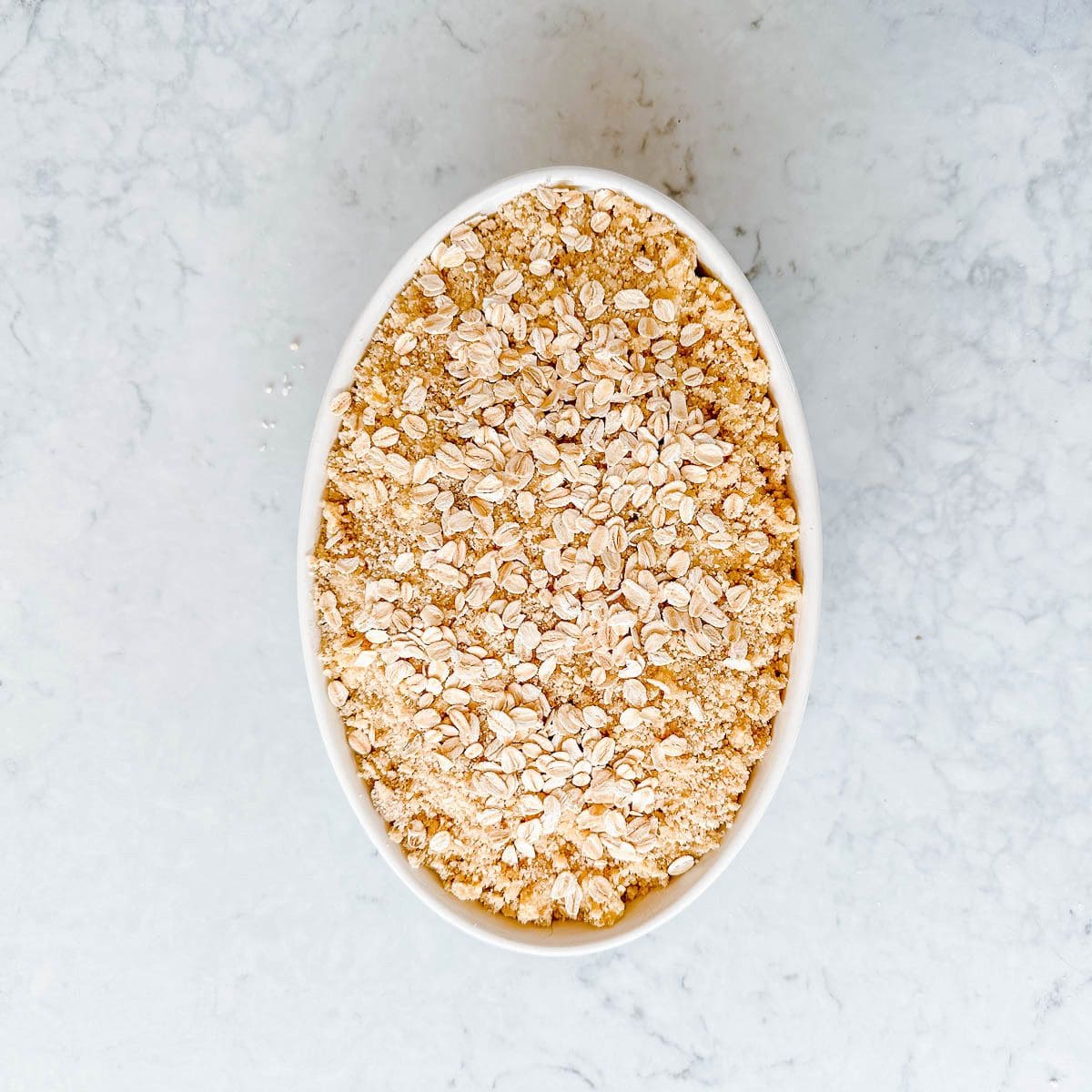 Oval white baking dish filled with an unbaked crumble dessert, set on a light grey marble surface.