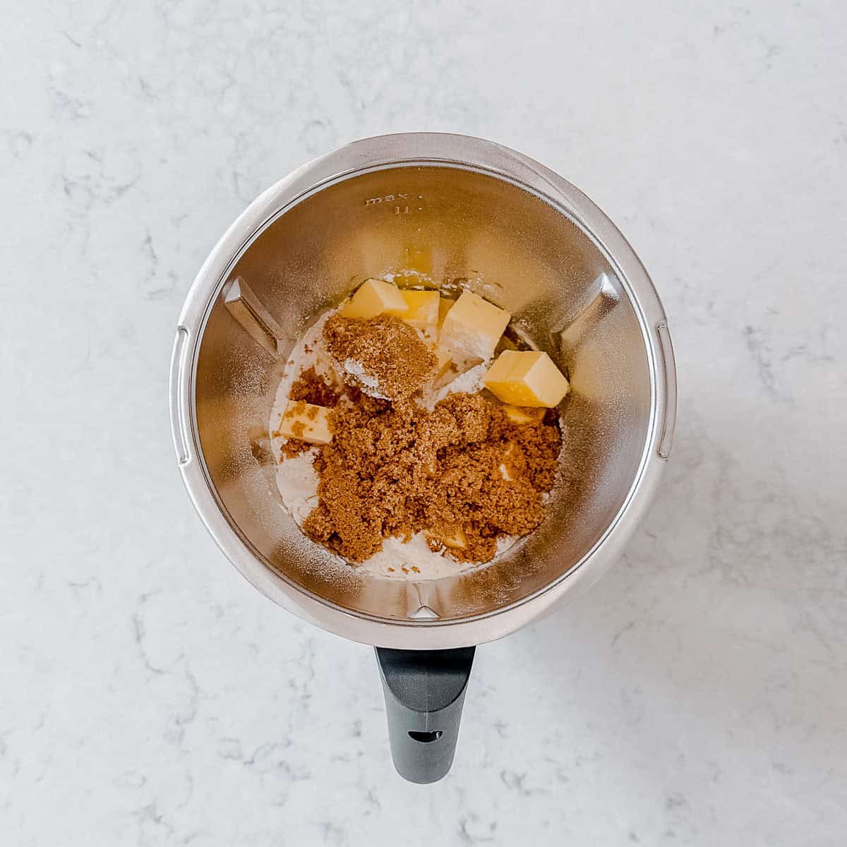 A top-down view of a Thermomix mixing bowl containing brown sugar, cubed butter, and other baking ingredients on a light marbled worktop.