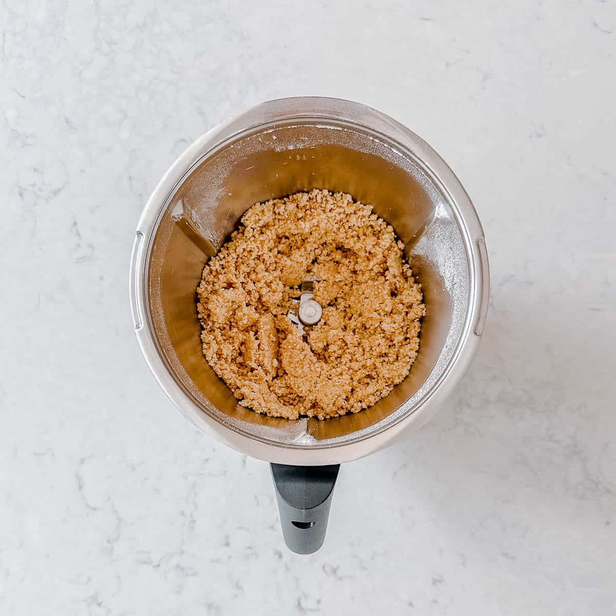 Overhead view of a Thermomix bowl containing a crumble mixture on a white marble worktop.