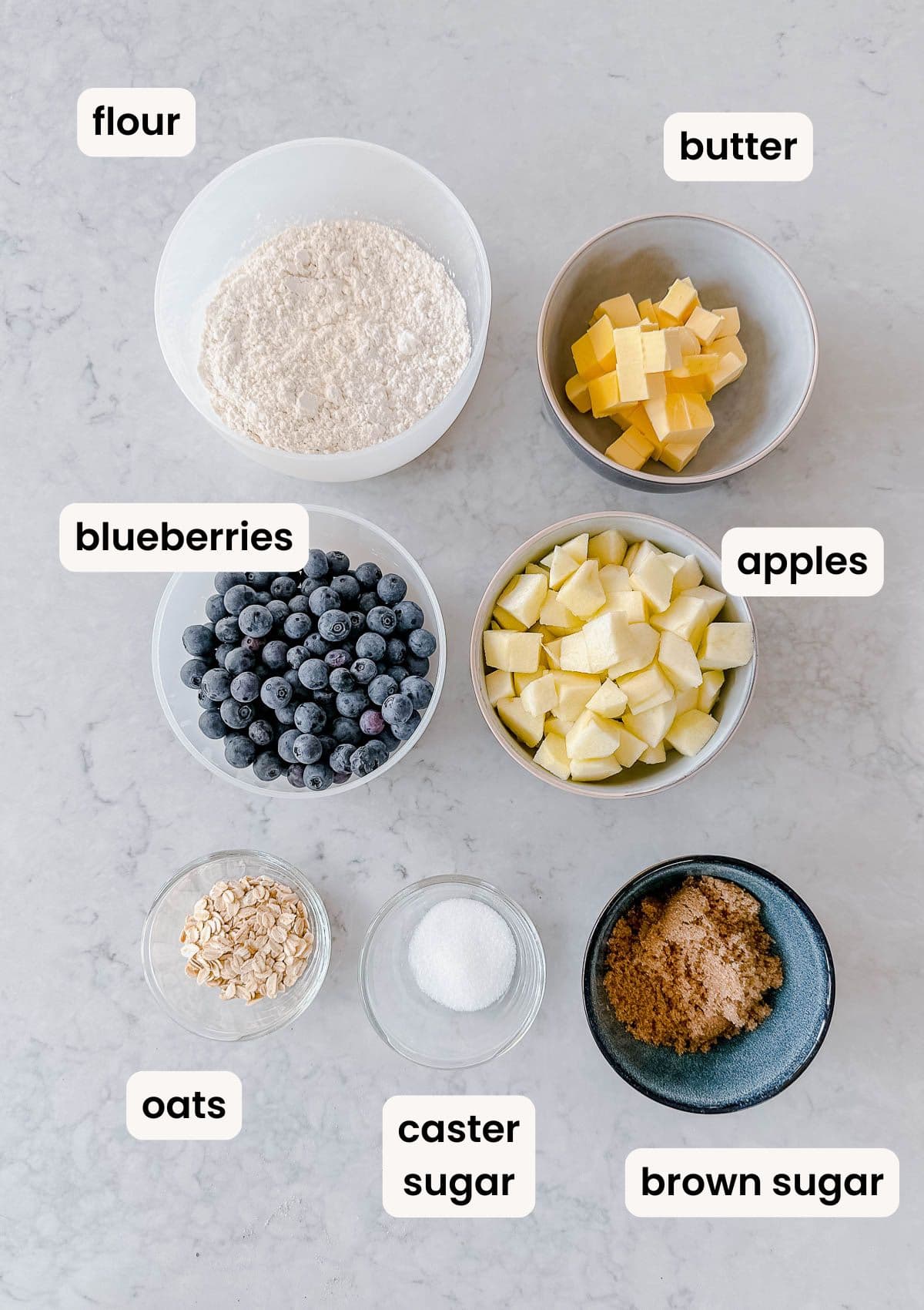Top-down view of bowls containing flour, butter, chopped apples, blueberries, oats, caster sugar, and brown sugar, each labelled with text on a light worktop.
