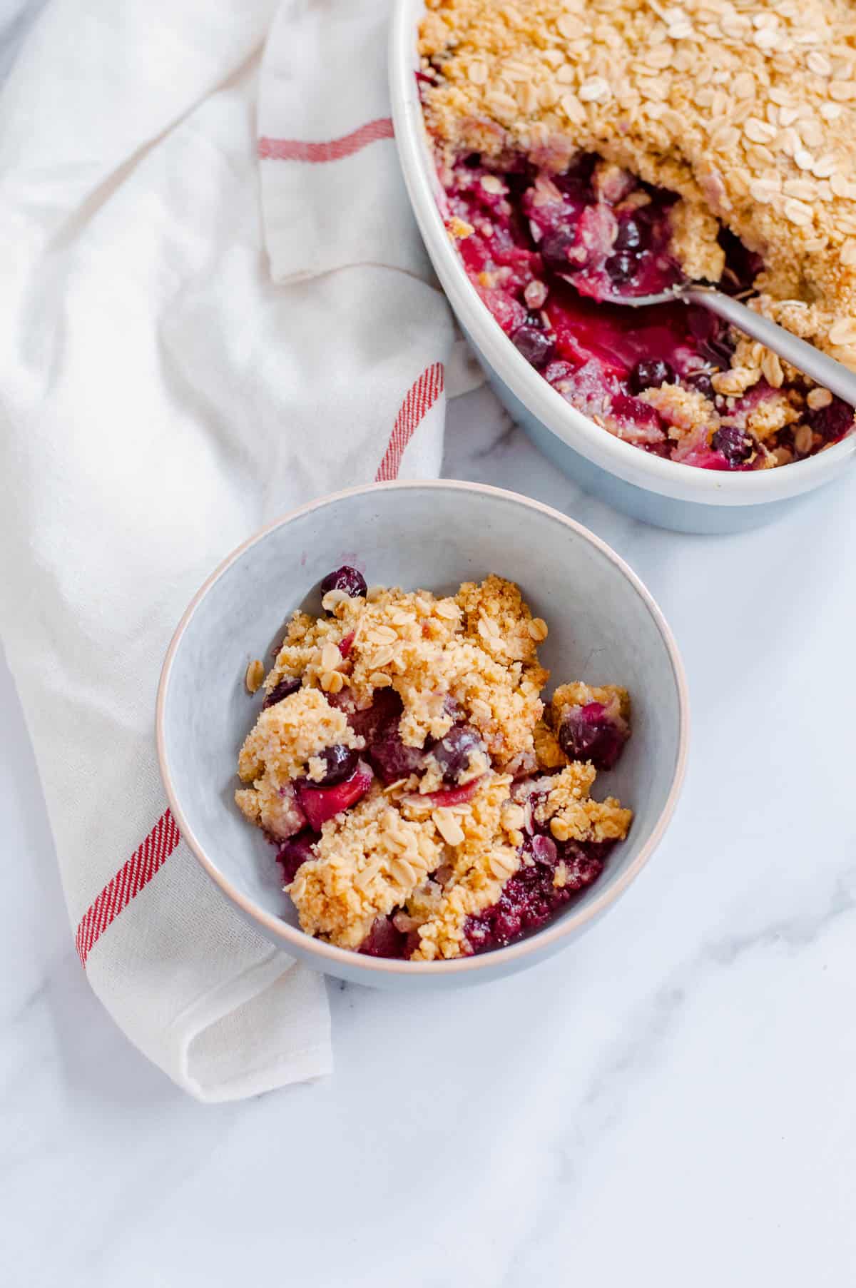A bowl of apple blueberry crumble with oats, positioned on a white surface next to a white serviette with a red stripe. Another dish of the crumble is partially visible in the background.