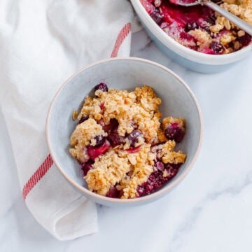 A bowl of apple blueberry crumble with oats, positioned on a white surface next to a white serviette with a red stripe. Another dish of the crumble is partially visible in the background.