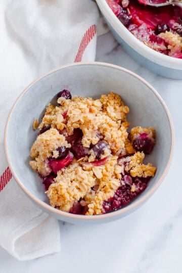 A bowl of apple blueberry crumble with oats, positioned on a white surface next to a white serviette with a red stripe. Another dish of the crumble is partially visible in the background.