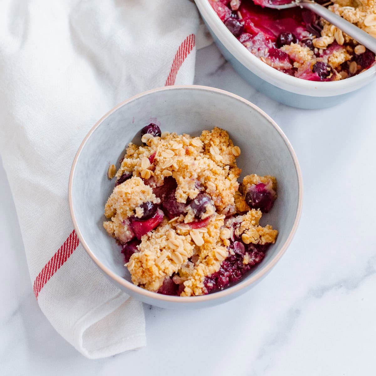 A bowl of apple blueberry crumble with oats, positioned on a white surface next to a white serviette with a red stripe. Another dish of the crumble is partially visible in the background.