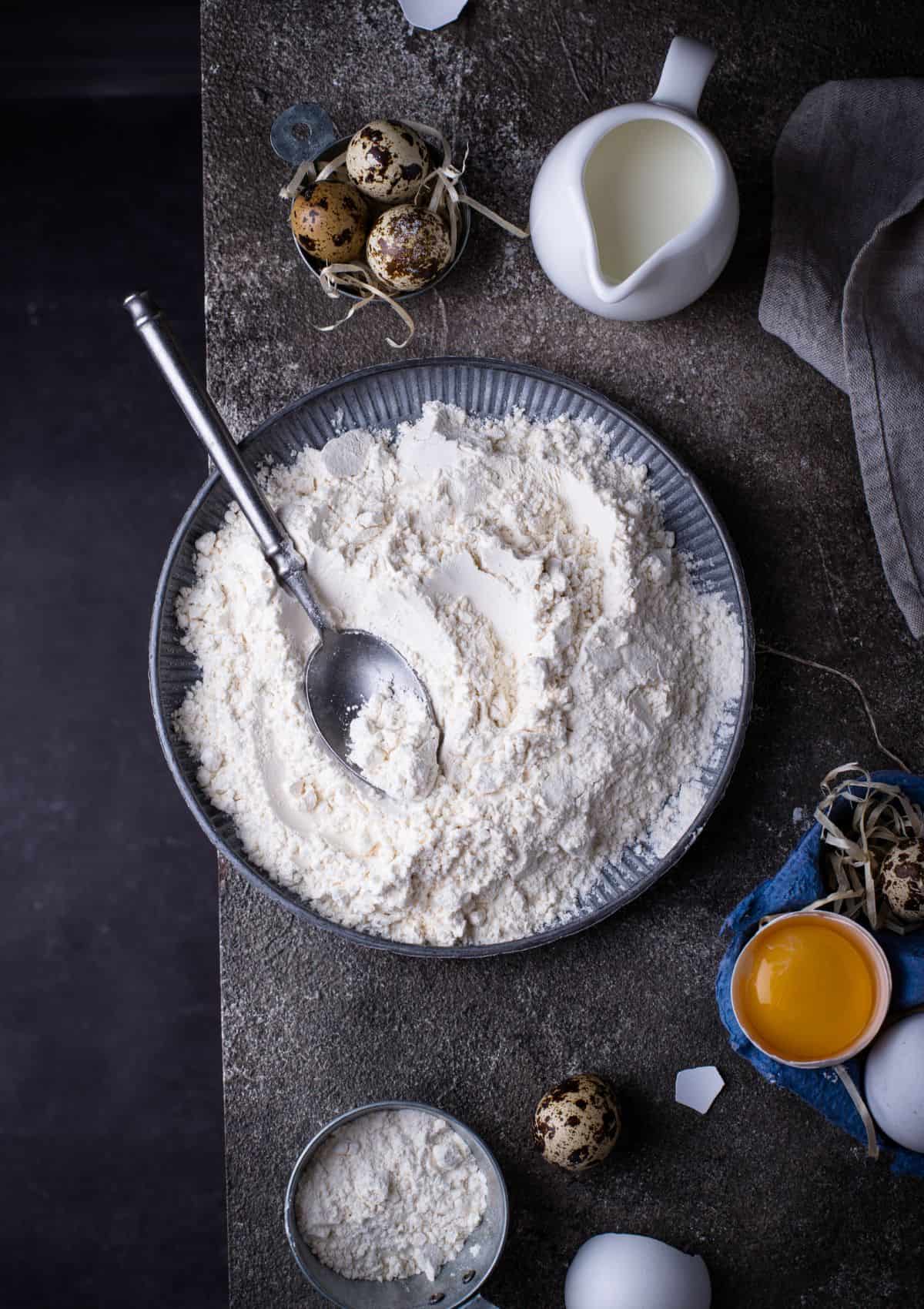 A bowl of flour with a metal spoon on a dark background.