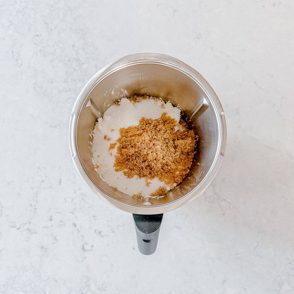 A Thermomix mixing bowl containing white granulated sugar and light brown sugar sits on a light-coloured, marbled worktop.