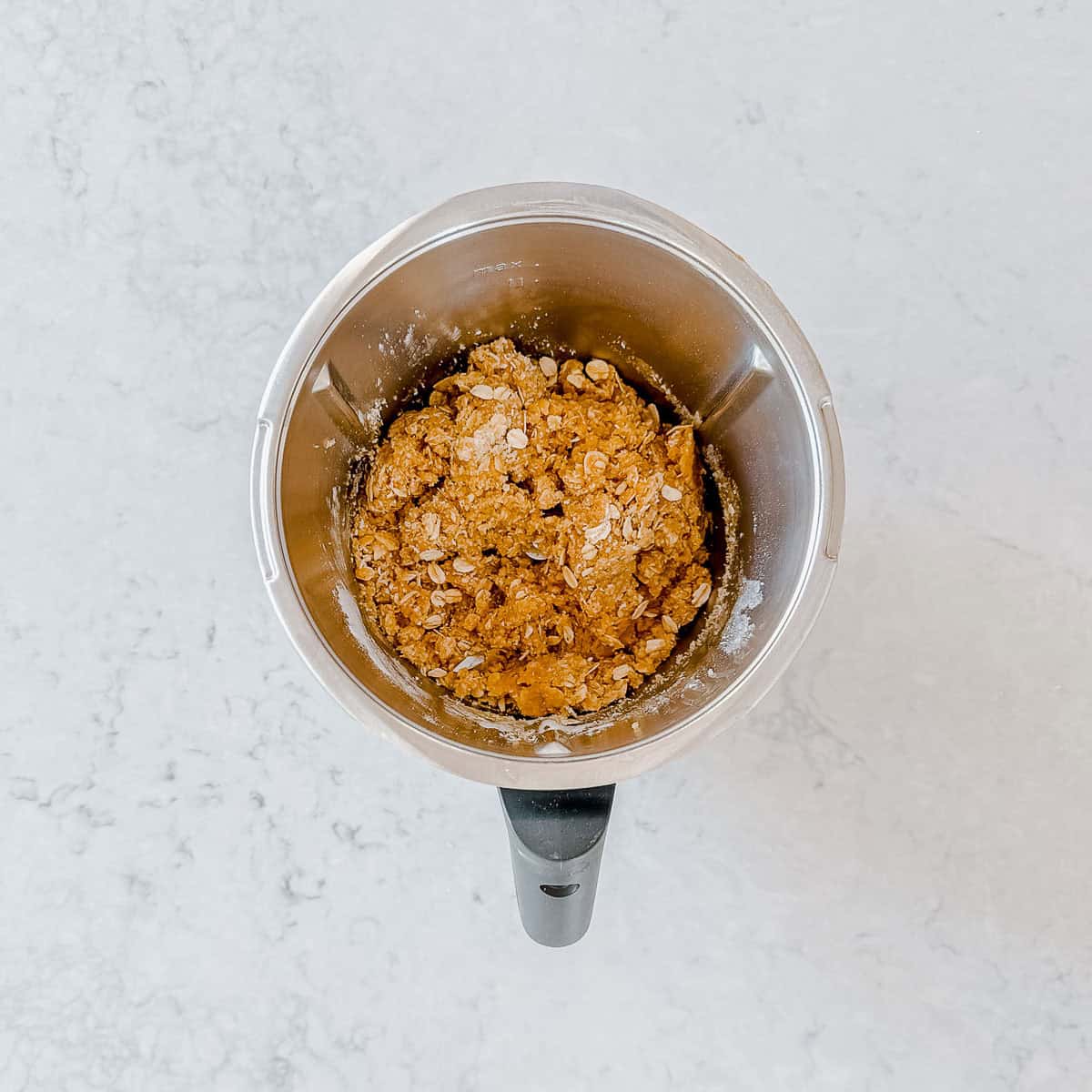 A top-down view of a thermomix bowl containing a thick oat-based dough on a light marble worktop.