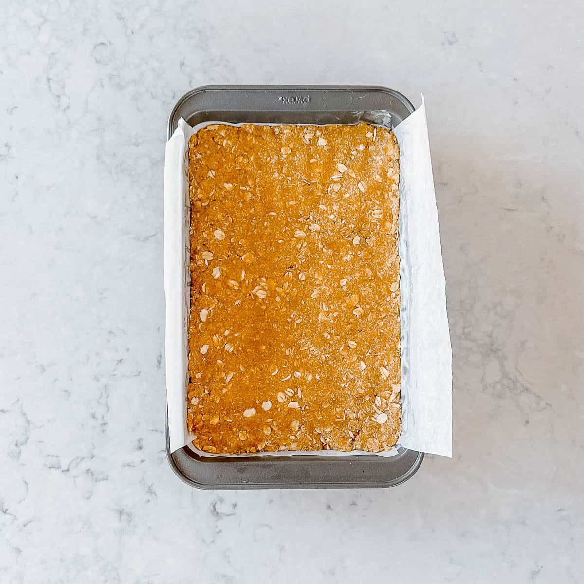 A rectangular metal baking tin lined with greaseproof paper holds an unbaked Anzac Slice, pressed evenly into the tin, on a light marble worktop.