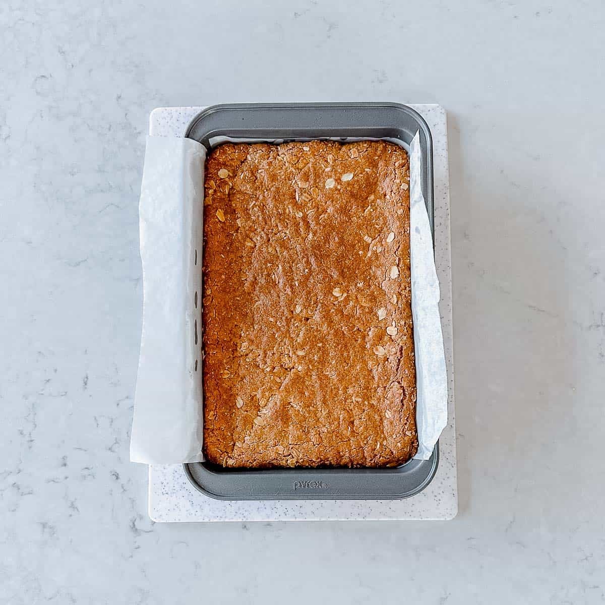A rectangular baking tin lined with greaseproof paper holds a large, golden-brown baked Anzac Slice, resting on a light-coloured worktop.