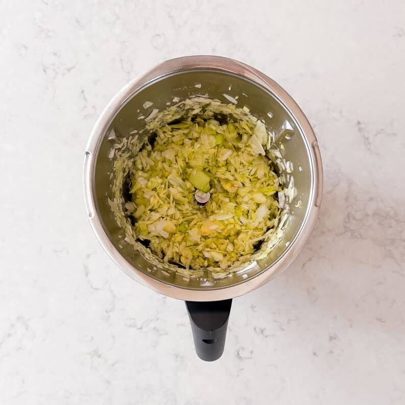 A top-down view of a food processor containing finely chopped leeks on a light marble worktop.
