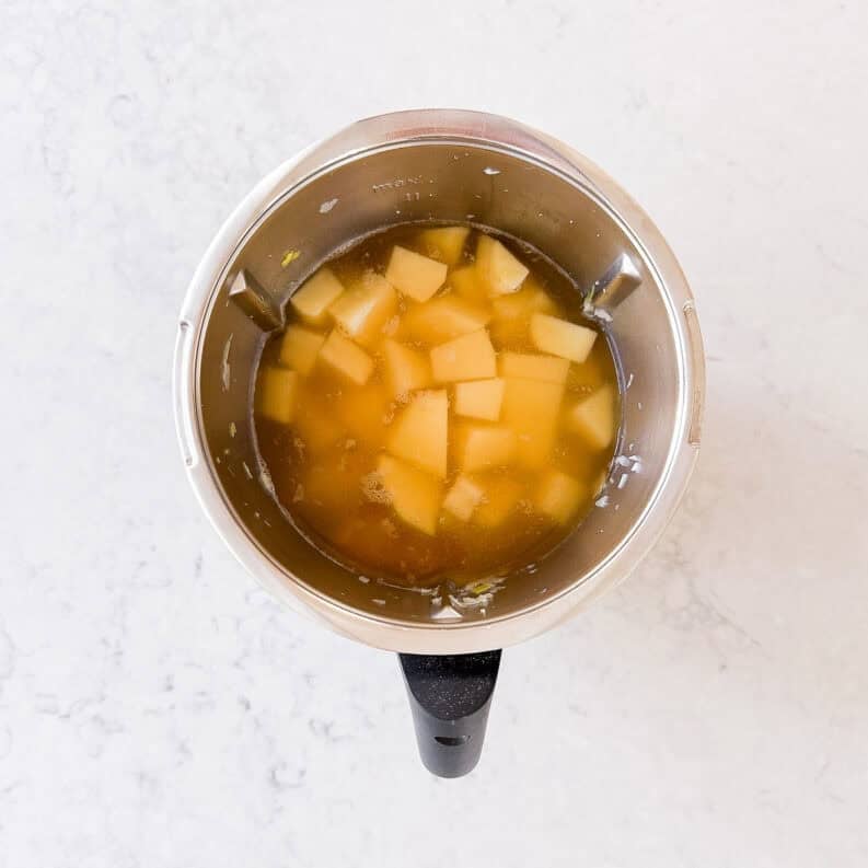 A top-down view of a metal saucepan containing cubed potatoes in stock, placed on a light-coloured, marble-like surface.