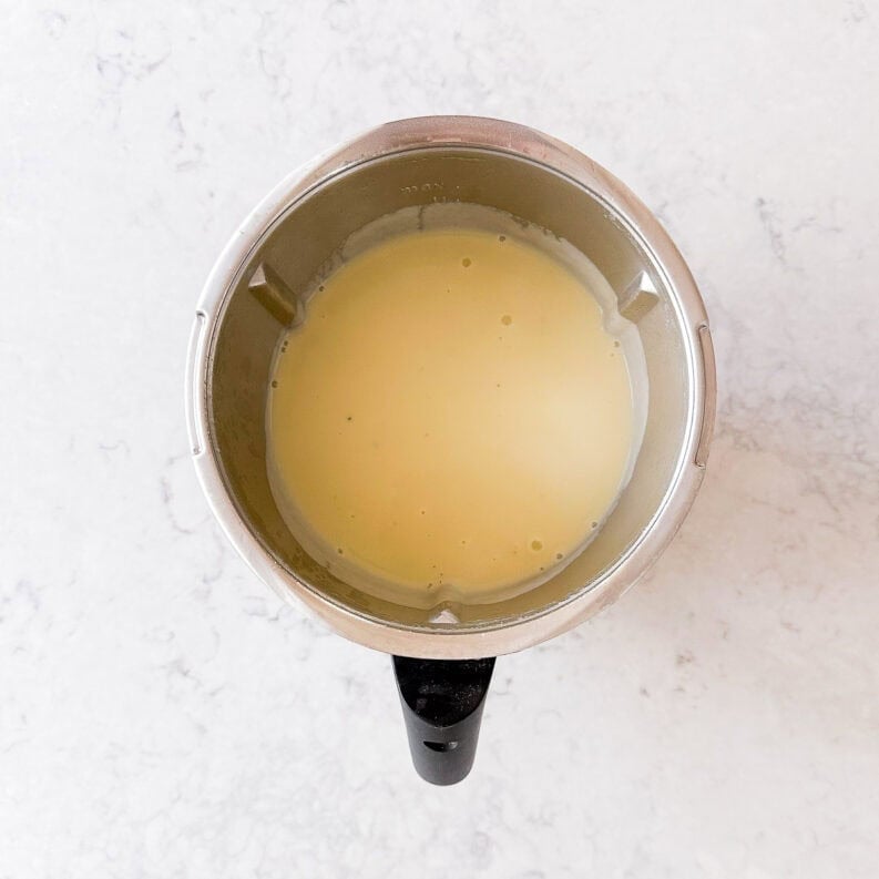 A top-down view of a blender containing a smooth, pale yellow liquid mixture, placed on a light marble worktop.