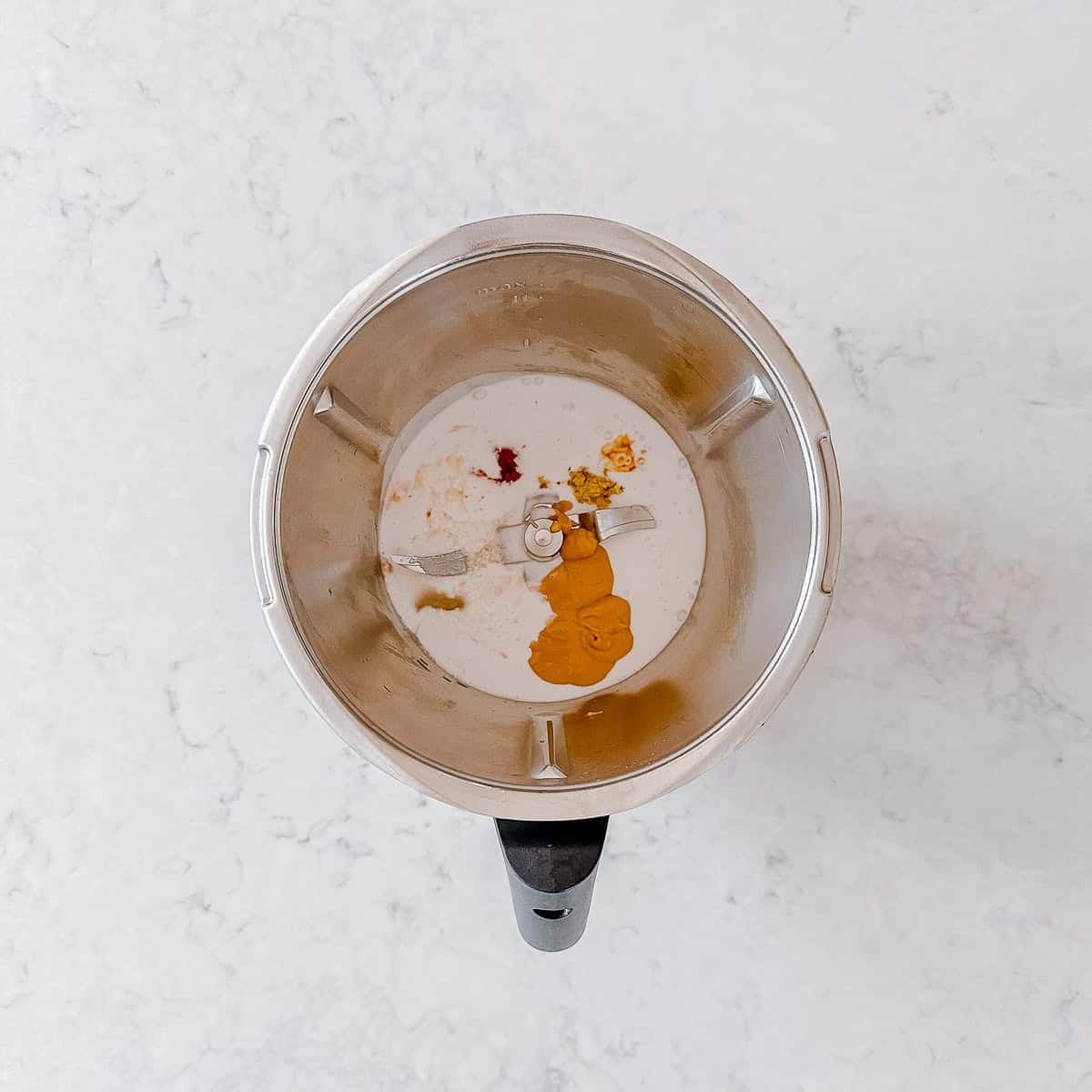 A top-down view of a blender containing a white liquid, likely milk, with dollops of orange, yellow, and red spices or pastes on a light-coloured worktop.