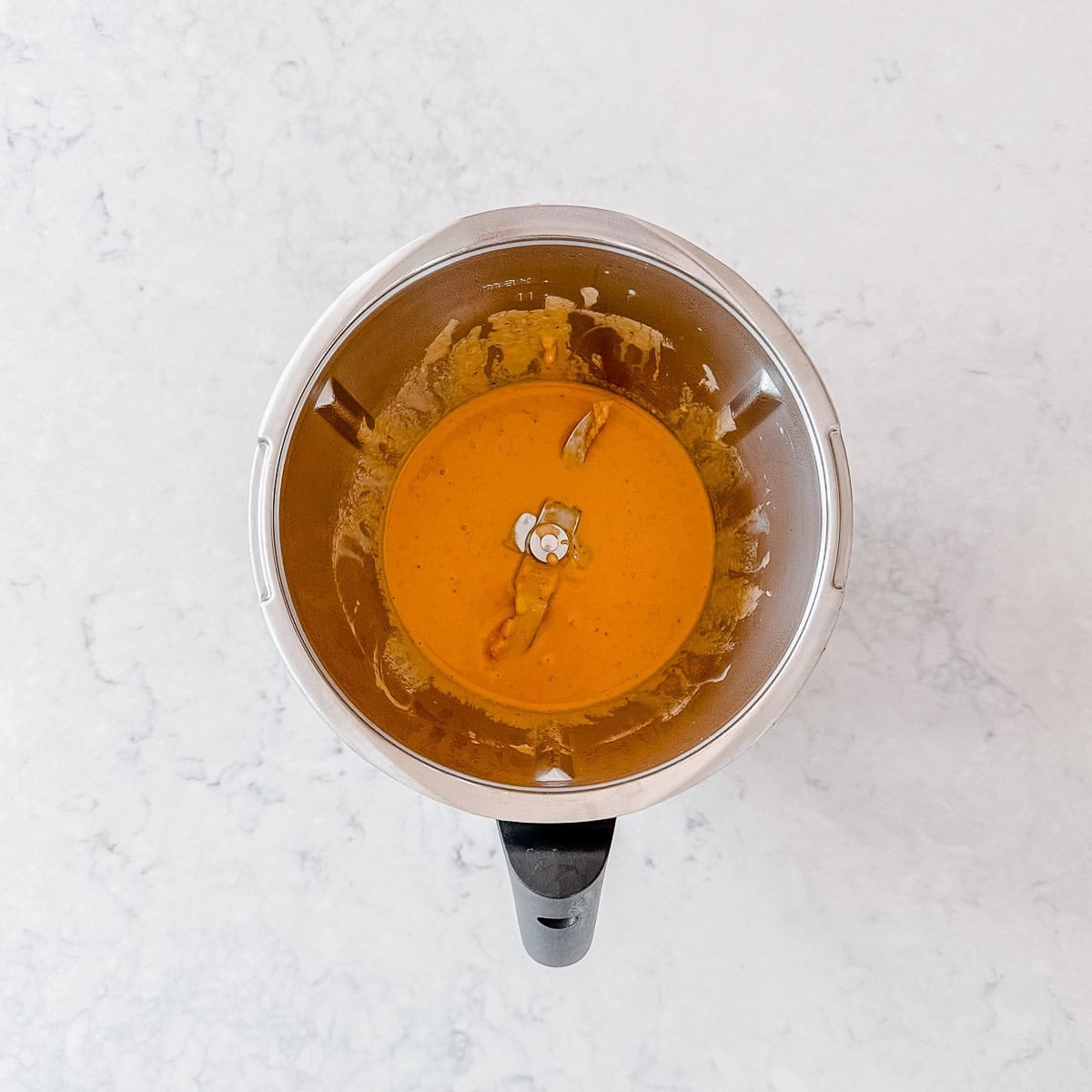A top-down view of a blender containing a smooth orange sauce with a small garlic clove and piece of pepper on top, placed on a light-coloured, textured worktop.