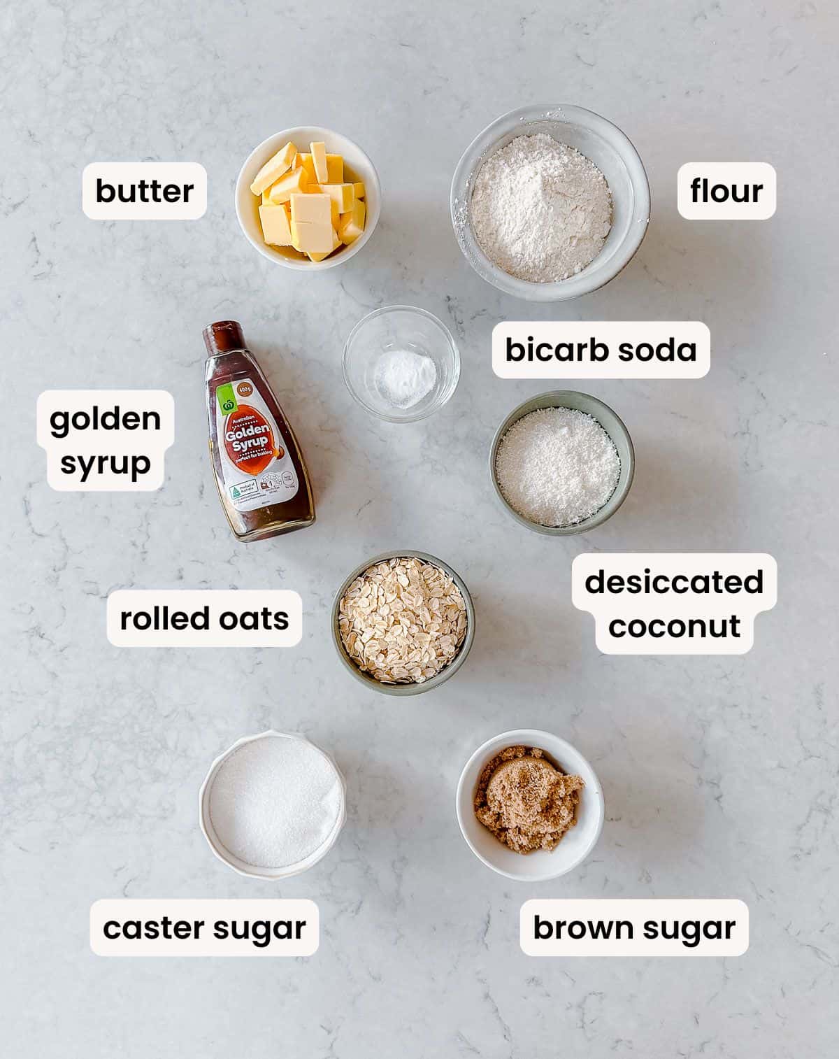 A flat lay of labelled baking ingredients on a white surface: butter, flour, golden syrup, bicarb soda, desiccated coconut, rolled oats, caster sugar, and brown sugar, each in separate bowls or containers.