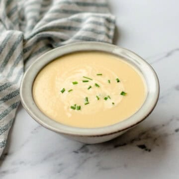 A bowl of creamy soup garnished with chopped chives sits on a white marble surface next to a striped cloth serviette.