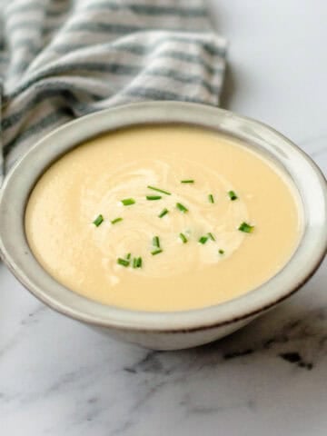 A bowl of creamy soup garnished with chopped chives sits on a white marble surface next to a striped cloth serviette.