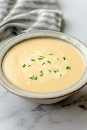 A bowl of creamy soup garnished with chopped chives sits on a white marble surface next to a striped cloth serviette.