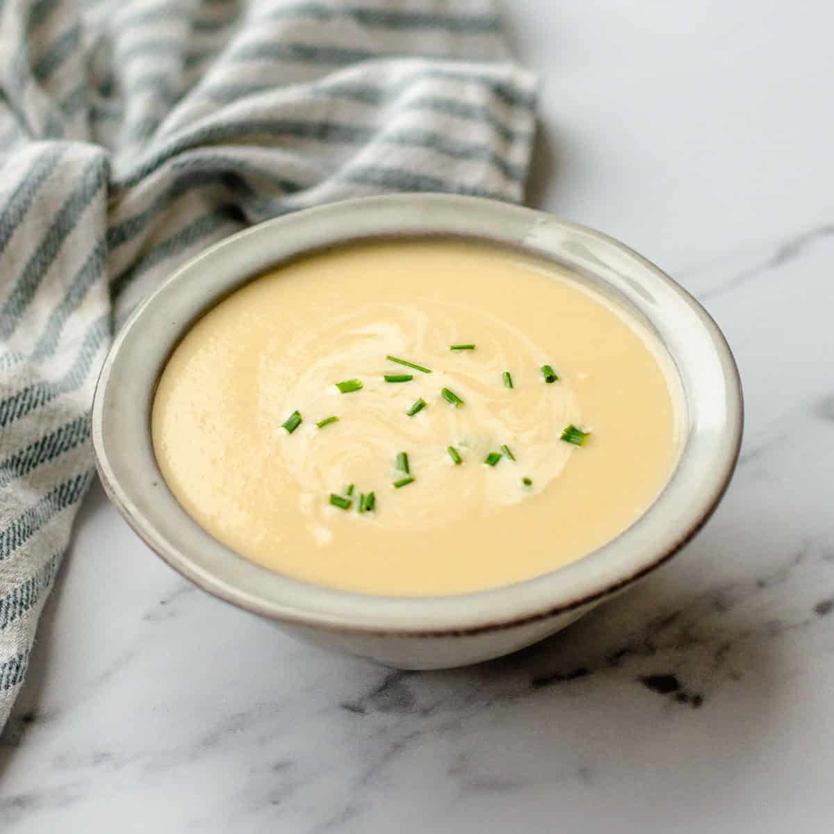 A bowl of creamy soup garnished with chopped chives sits on a white marble surface next to a striped cloth serviette.