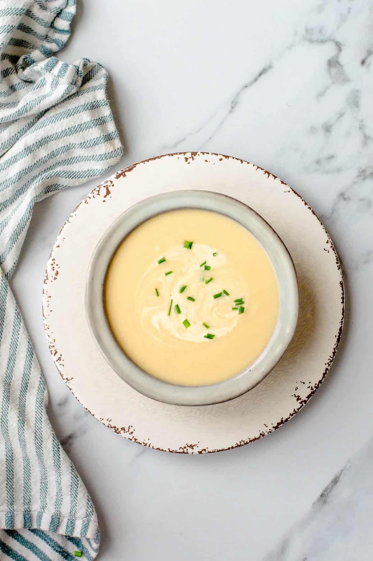 Overhead view of a creamy Thermomix leek and potato soup in a light grey bowl on a rustic white plate, topped with a drizzle of cream and chopped fresh chives.
