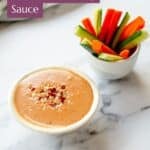 A bowl of satay sauce garnished with chopped nuts and red pepper flakes sits beside a cup filled with carrot and cucumber sticks on a white marble surface.