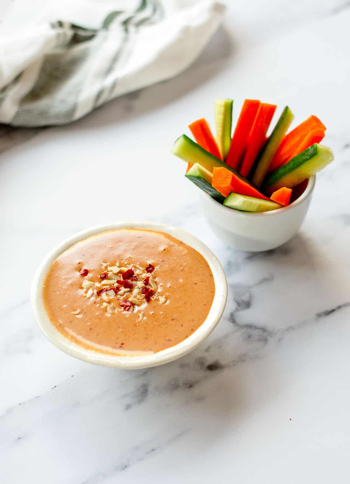A bowl of satay sauce garnished with chopped nuts and red pepper flakes sits beside a cup filled with carrot and cucumber sticks on a white marble surface.