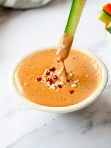 A cucumber spear is dipped into a bowl of creamy, peanut satay sauce, garnished with chopped peanuts and red pepper flakes.