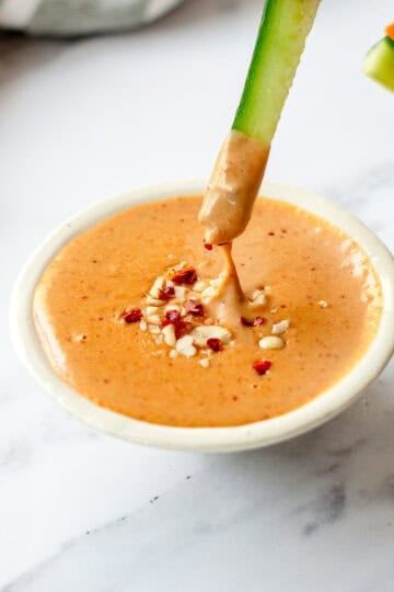A cucumber spear is dipped into a bowl of creamy, peanut satay sauce, garnished with chopped peanuts and red pepper flakes.