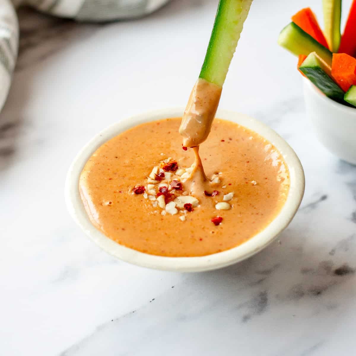 A cucumber spear is dipped into a bowl of creamy, peanut satay sauce, garnished with chopped peanuts and red pepper flakes.