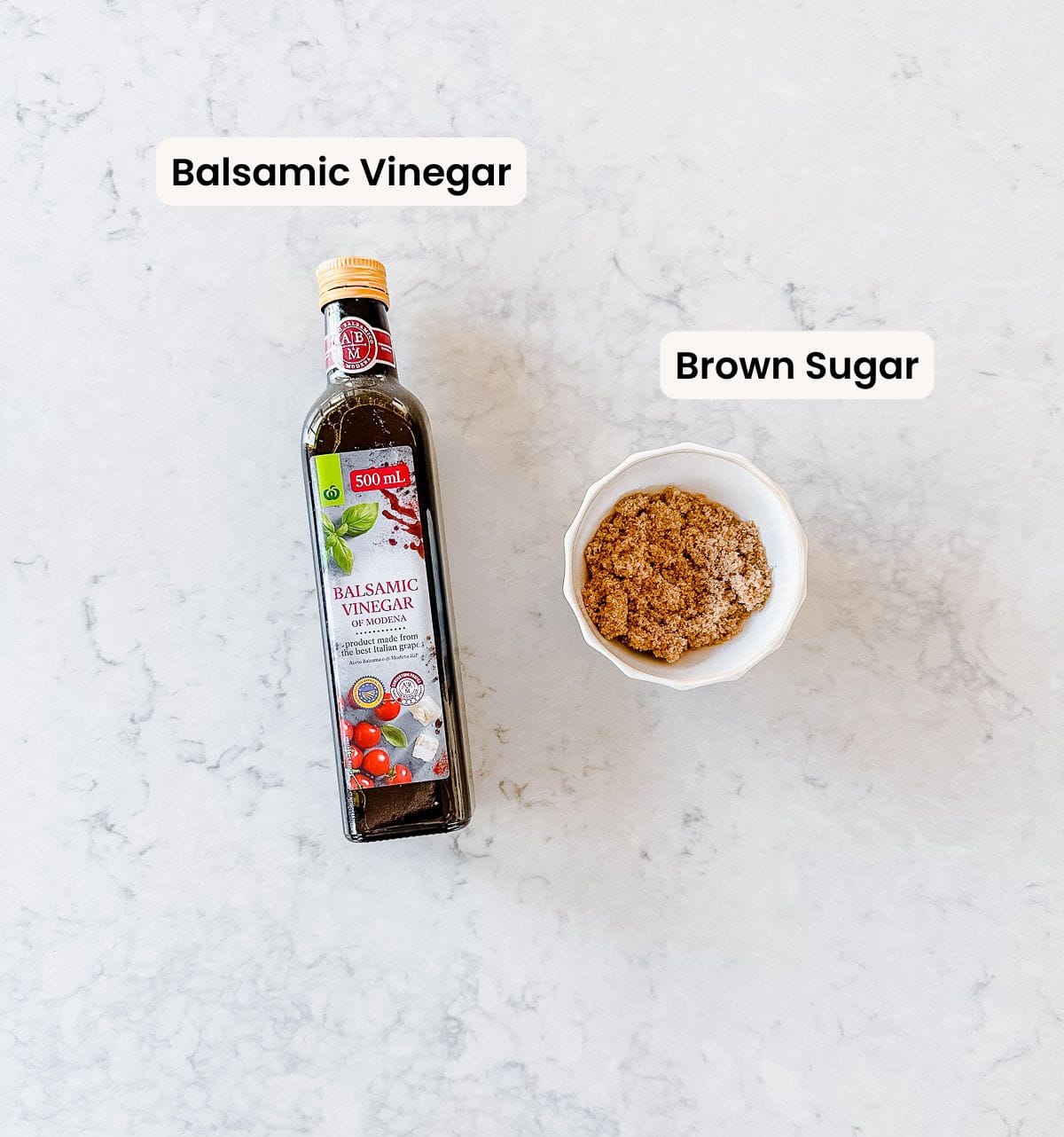 A bottle of balsamic vinegar, and a bowl of brown sugar, labelled and sitting on a light grey marble benchtop.
