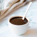 A small white bowl filled with balsamic glaze, with a white teaspoon sticking out. On a white marble background with a light coloured tea towel in the distance.