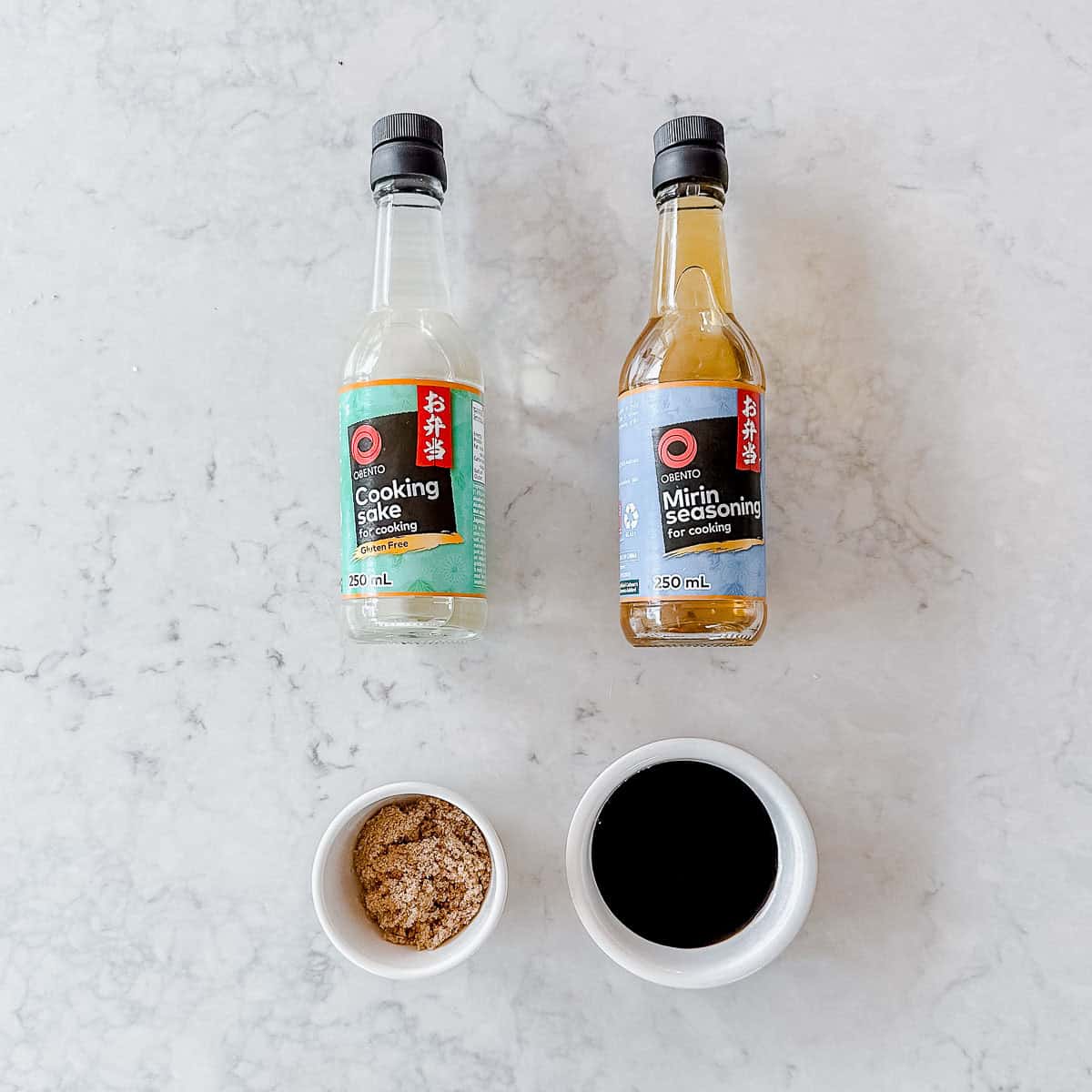 Two bottles of Obento cooking sake and mirin seasoning, with small bowls of brown sugar and soy sauce beneath them.
